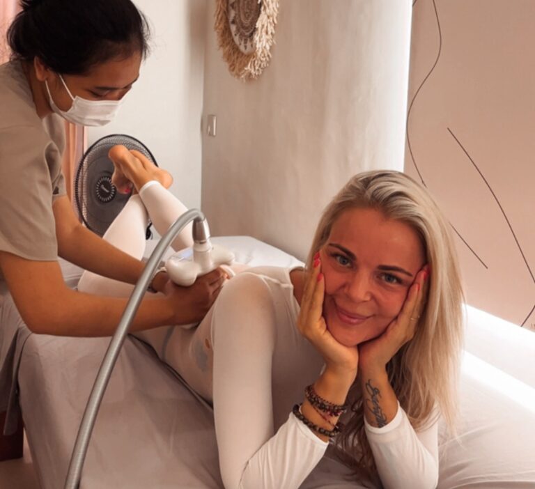 A therapist performs an LPG endermologie treatment on a client's upper arm through a white treatment suit.