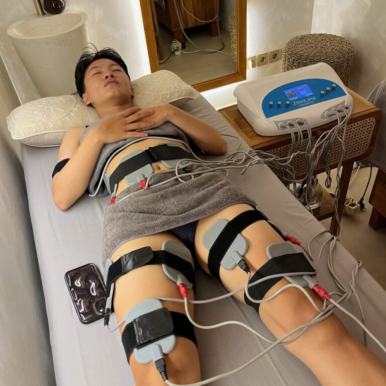 A client lies on a treatment bed with electrical stimulation pads secured to the abdomen and thighs.