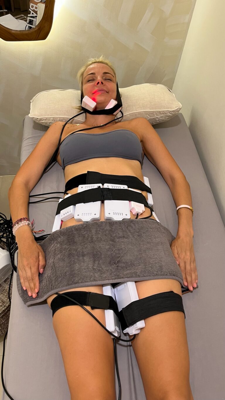 A client receives cold lipo laser treatment with red-lit pads secured to her chin, abdomen, and thighs.