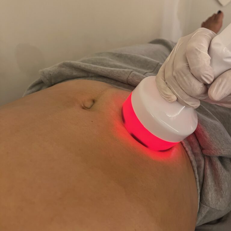 A therapist applies a red-lit handheld probe to a client's abdomen during a body treatment.