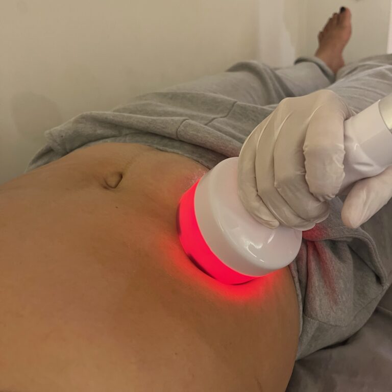 A therapist applies a red-lit handheld probe to a client’s abdomen during a body treatment.